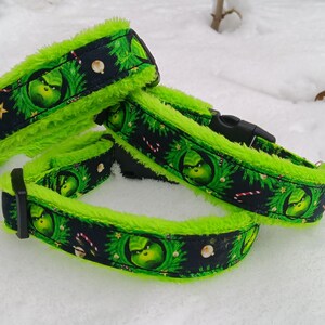Könnte beinhalten: Drei Hundehalsbänder mit einem Grinch-Motiv. Die Halsbänder haben einen schwarzen Hintergrund mit Grinch-Gesichtern, Zuckerstangen und Sternen. Die Ränder sind mit leuchtend grünem, flauschigem Material gefüttert. Die Halsbänder liegen im Schnee.