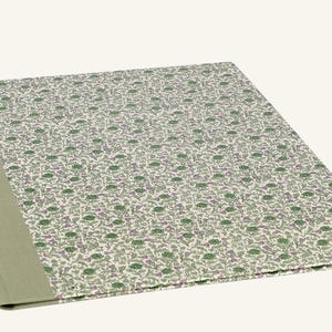 Op de afbeelding: Een gesloten, rechthoekig notitieboek met een bloemenpatroon op de kaft. De kaft heeft een herhalend ontwerp van groene en paarse bloemen op een crèmekleurige achtergrond. De rug is effen, gedempt groen.