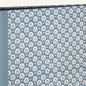 Könnte beinhalten: Nahaufnahme eines gemusterten Buches oder einer Schachtel. Der Einband zeigt ein sich wiederholendes Blumendesign in Weiß und Pfirsich auf blauem Hintergrund. Der Buchrücken ist einfarbig blau. Die Gesamtästhetik ist Vintage oder Bohemian.