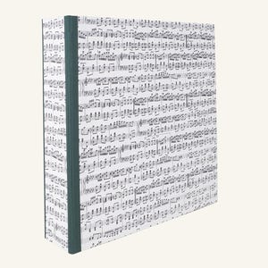 May include: A hardcover notebook with a musical score pattern. The cover is white with black musical notes, and the spine is a dark teal color. The notebook is rectangular and stands upright.