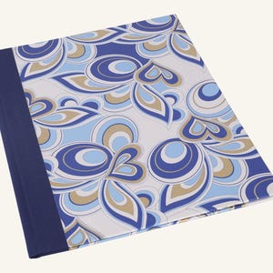 May include: A hardcover notebook with a navy blue spine and a patterned cover. The cover features a repeating design of blue, light blue, and tan abstract shapes on a white background. The notebook is angled slightly.