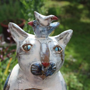 Könnte beinhalten: Eine Keramikkatzenfigur mit weiß-grau gesprenkelter Glasur, leuchtend blauen Augen, einer kleinen roten Nase und einem kleinen Vogel auf dem Kopf. Der Vogel ist weiß mit blauen Flügeln und einem roten Schnabel und setzt einen farbigen Akzent.