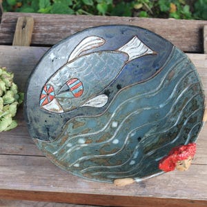Könnte beinhalten: Ein handgefertigter Keramikteller mit Fischmotiv. Der Teller ist überwiegend blau, mit einem Fischdesign in der Mitte, das weiße, rote und blaue Akzente aufweist. Er hat ein wellenförmiges Muster und eine kleine Meerjungfrau.