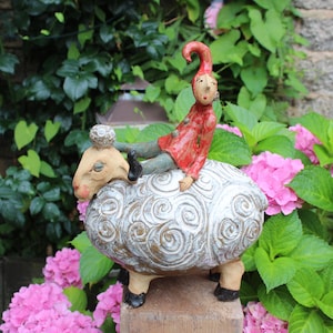 Könnte beinhalten: Eine skurrile Keramikskulptur eines Schafs mit einer sitzenden Figur. Das Schaf ist weiß mit einem strukturierten Muster, die Figur trägt einen roten Mantel und Hut. Die Skulptur steht vor rosa Blüten und grünem Laub.