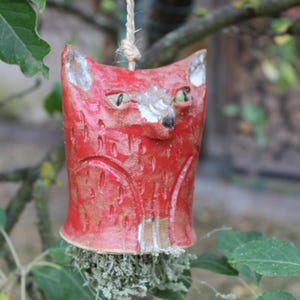 Könnte beinhalten: Ein rotes Keramik-Fuchs-Ornament mit spitzen Ohren, grünen Augen und einer schwarzen Nase, das an einer hellen Schnur hängt. Der Fuchs hat eine strukturierte Oberfläche mit eingearbeiteten Details und ein Büschel getrocknetes grünes Moos.