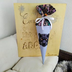 Könnte beinhalten: Hellblauer, kegelförmiger Geschenkebeutel mit einem schwarzen Panthergesicht. Der Beutel ist mit einem gemusterten Stoff und weißem Band gebunden. Im Hintergrund befindet sich eine Holztafel mit den Buchstaben "ABC" und dem Wort "Einschulung".