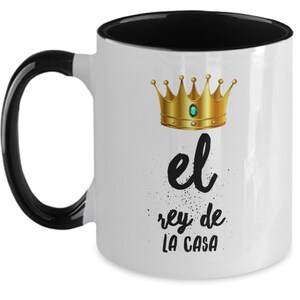 Puede incluir: Taza de cer&aacute;mica blanca con interior y asa negros. La taza presenta una corona dorada con una gema verde y el texto en espa&ntilde;ol "el rey de la casa" en escritura negra.