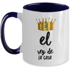 Puede incluir: Taza de cer&aacute;mica blanca con interior y asa azul marino. La taza presenta una corona dorada con una gema verde y el texto "el rey de LA CASA" en escritura negra.