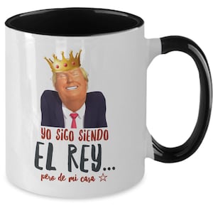 May include: White ceramic mug with a black interior and handle. The mug features a smiling caricature wearing a gold crown. Text in Spanish reads "Yo sigo siendo El Rey... pero de mi casa" in black and red.