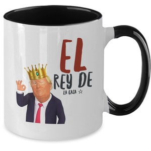 Könnte beinhalten: Weißer Keramikbecher mit schwarzem Innenraum und Henkel. Der Becher zeigt eine Illustration eines Mannes mit goldener Krone und dem Text "EL REY DE LA CASA". Der Mann macht die "Okay"-Geste.