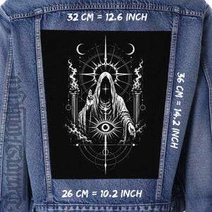 Pode incluir: Um casaco de ganga azul com um grande emblema retangular preto nas costas. O emblema apresenta um gráfico branco de uma figura encapuzada, um olho e símbolos celestiais. O texto indica dimensões de 32 cm por 36 cm.