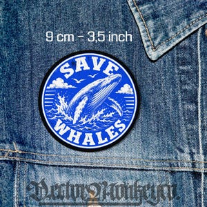 May include: A circular embroidered patch measuring 9 cm in diameter. The patch features a blue background with a white illustration of a breaching whale, ocean waves, clouds, and birds. The text SAVE WHALES is printed in white.