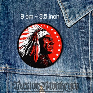 May include: Round patch featuring a Native American chief in profile, rendered in red, black, and white. The patch is 3.5 inches (9 cm) in diameter and is set against a red background with white dots. The patch is on a denim jacket.