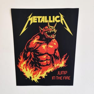 May include: Black graphic featuring the band Metallica's logo in yellow, above a red and orange demon illustration. The text "JUMP IN THE FIRE" is printed in red at the bottom. The image is on a black background.
