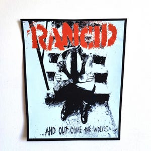 Puede incluir: Un parche cuadrado con borde negro. Presenta una ilustración en blanco y negro de una persona sentada en unos escalones con los brazos cruzados. La palabra "RANCID" está en letras grandes de color naranja en la parte superior, y el texto "...AND OUT COME THE WOLVES" en la parte inferior.