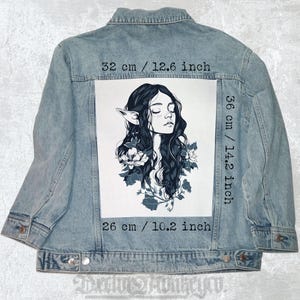 May include: Pale blue denim jacket with a large white graphic on the back. The graphic depicts a black and white illustration of a woman with long hair, pointed ears, and flowers. The graphic measures 32 cm wide, 36 cm tall, and 26 cm long.