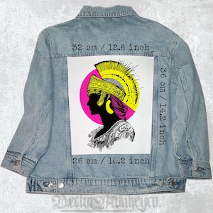 Pode incluir: Uma jaqueta de ganga com um remendo nas costas. O remendo é branco com uma ilustração em preto e branco de um perfil de mulher usando um capacete amarelo e dourado. O remendo mede 36 cm por 26 cm.