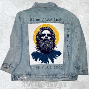 Pode incluir: Uma jaqueta de ganga azul com um remendo branco nas costas. O remendo apresenta uma ilustração em preto e branco de um homem com barba e cabelo comprido. O homem está rodeado por um círculo amarelo. O remendo tem 36 cm de altura e 26 cm de largura.