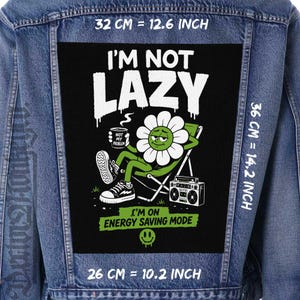 Puede incluir: Una chaqueta vaquera azul con un gran parche rectangular en la espalda sobre fondo negro. El gráfico muestra una flor blanca en una silla de playa con un radiocasete, texto I'M NOT LAZY y I'M ON ENERGY SAVING MODE. Dimensiones de 32 cm por 36 cm.