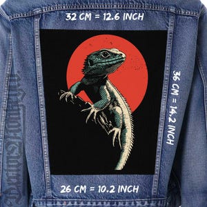 Op de afbeelding: Een blauwe spijkerjas met een grote, rechthoekige patch op de rug. De patch toont een gedetailleerde illustratie van een hagedis in blauw en wit, tegen een rode cirkel op een zwarte achtergrond. De patch meet 32 cm bij 36 cm.