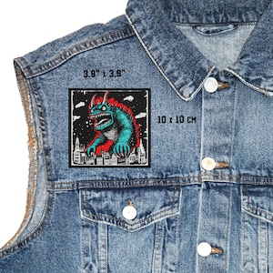May include: A square patch featuring a monster design, measuring 10 x 10 cm. The monster is teal and red, with a city skyline at the bottom. The patch is on a denim jacket.