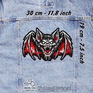 Peut inclure: Écusson de chauve-souris noir et rouge, 30 cm de large et 19 cm de haut, avec une bouche ouverte et des dents pointues. L'écusson est thermocollant et est affiché sur une veste en jean.