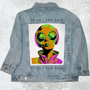 Op de afbeelding: Lichtblauwe spijkerjas met een opvallende alien-afbeelding op de rug. De afbeelding, 32 x 36 x 26 cm, toont een alien met grote ogen in groen, oranje en paars. De jas heeft een klassiek ontwerp.