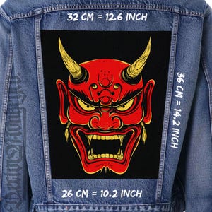 Peut inclure: Veste en jean avec un grand patch rectangulaire représentant un visage de démon rouge avec des cornes et des yeux jaunes. Le patch mesure 32 cm de large, 36 cm de haut et 26 cm de large. Le texte "32 CM = 12.6 INCH", "36 CM = 14.2 INCH", et "26 CM = 10.2 INCH" est visible.