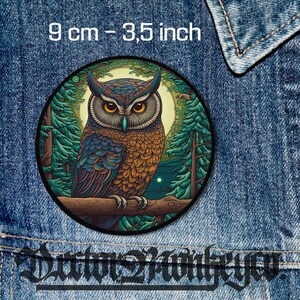 Könnte beinhalten: Ein runder Patch mit einer detaillierten Illustration einer Eule, die auf einem Ast in einer Waldlandschaft sitzt. Die Eule hat braune und blaue Federn, gelbe Augen und einen dunklen Schnabel. Der Hintergrund zeigt grüne Bäume und einen Mond am Himmel. Der Patch misst 9 cm oder 3,5 Zoll im Durchmesser.