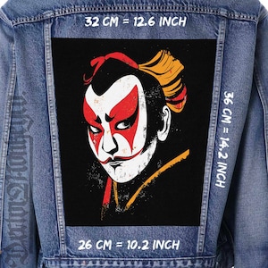 May include: A blue denim jacket featuring a large rectangular patch on the back with a graphic of a face in white, red, and yellow makeup on a black background. Text indicates dimensions of 32 cm = 12.6 inches, 36 cm = 14.2 inches, and 26 cm = 10.2 inches.