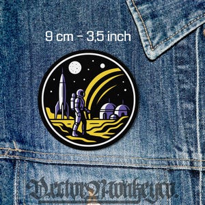 Peut inclure: Un écusson rond sur le thème de l'espace, avec une fusée, un astronaute et des structures extraterrestres sur fond de ciel noir étoilé. Le patch mesure 9 cm (3,5 pouces) de diamètre. Le design comprend des accents jaunes et blancs.