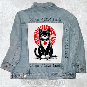 Peut inclure: Veste en jean bleu clair avec un grand motif au dos. Le motif représente un chat noir avec un motif de soleil rouge. Le motif mesure 32 cm de large, 36 cm de haut et 26 cm en bas.