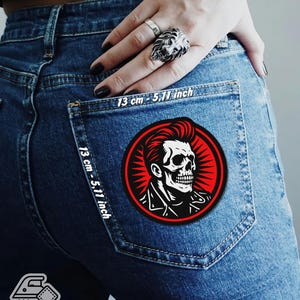 May include: A round, iron-on patch featuring a stylized skull with a pompadour hairstyle, set against a red burst design. The patch is 5.11 inches (13 cm) in diameter and is displayed on a pair of blue jeans. The patch has the text "Iron On Patch".