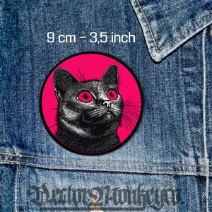 May include: A black and white illustration of a cat with bright red eyes, set against a pink circle. The image is 3.5 inches in diameter.