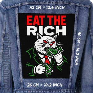 Peut inclure: Veste en jean avec un grand écusson. L'écusson présente un chat blanc en costume, aux yeux rouges et cravate rouge, mordant de l'argent, avec le texte "EAT THE RICH". Dimensions de l'écusson : 26 cm x 36 cm.