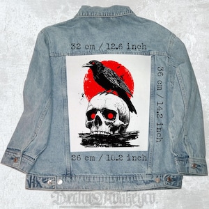 Könnte beinhalten: Hellblaue Jeansjacke mit einem Grafikdesign auf dem Rücken. Das Design zeigt einen schwarzen Raben auf einem Totenkopf mit roten Augen, vor einem roten Sonnenhintergrund. Das Design misst 32 cm breit, 36 cm hoch und 26 cm lang.