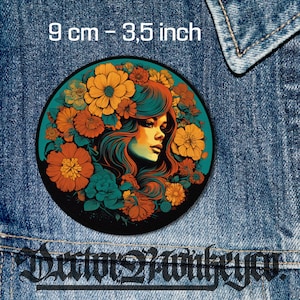 Könnte beinhalten: Ein runder Patch mit einer farbenfrohen Illustration einer Frau mit langen, fließenden Haaren, umgeben von Blumen. Die Blumen sind in Orange-, Gelb- und Rottönen gehalten. Der Hintergrund ist blaugrün. Der Patch hat einen Durchmesser von 9 cm.