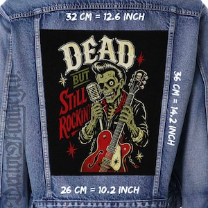 Könnte beinhalten: Jeansjacke mit einem rechteckigen Aufnäher, der ein Skelett zeigt, das eine rote Gitarre spielt und ein Mikrofon hält. Der Aufnäher trägt die Aufschrift "DEAD BUT STILL ROCKIN'" in weißen und roten Buchstaben. Der Aufnäher ist 32 cm breit, 36 cm hoch und 26 cm lang.