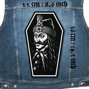 Può includere: Una toppa in bianco e nero raffigurante un uomo con baffi e cappello ornato, in una cornice a forma di bara. La toppa è alta 36 cm e larga 22 cm, applicata su un gilet di jeans.