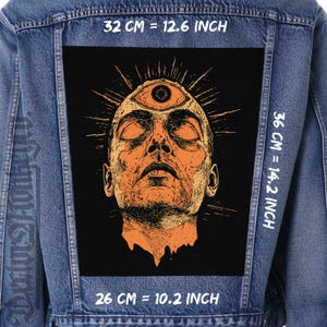 Pode incluir: Um remendo preto e laranja com uma ilustração estilizada de uma pessoa com um terceiro olho na testa. O remendo está em uma jaqueta de jeans azul. O texto "32 CM = 12.6 INCH", "36 CM = 14.2 INCH", e "26 CM = 10.2 INCH" está impresso na jaqueta.
