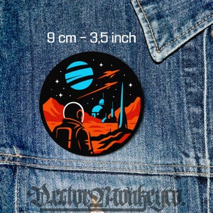 May include: A round, black patch with a space-themed design. It features an astronaut looking at a planet with blue stripes, rockets, and a futuristic city. The patch is 3.5 inches in diameter.