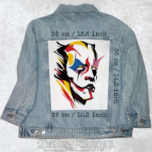 May include: A denim jacket with a colorful graphic patch on the back. The patch features a stylized face with red, yellow, blue, and pink accents. The patch measures 14.2 inches by 10.2 inches.