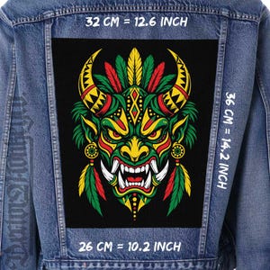 Puede incluir: Una chaqueta de mezclilla azul con un parche rectangular grande en la espalda que muestra un gráfico colorido de una máscara de demonio con cuernos en rojo, amarillo y verde. El texto indica dimensiones de 32 cm = 12.6 pulgadas, 36 cm = 14.2 pulgadas y 26 cm = 10.2 pulgadas.