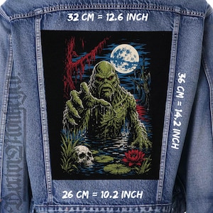 May include: A blue denim jacket featuring a large rectangular back patch with a swamp creature illustration. The graphic shows a green monster in water with a skull, a red flower, and a full moon. Dimensions are 32 cm by 36 cm.