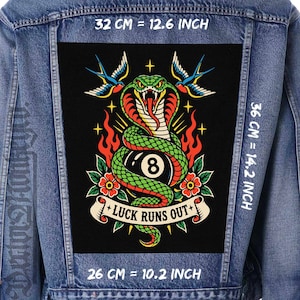 Puede incluir: Una chaqueta de mezclilla azul con un parche rectangular grande en la espalda que muestra una cobra verde enroscada alrededor de una bola de billar negra número 8, flanqueada por golondrinas, llamas y flores, con el texto LUCK RUNS OUT. Mide 32 cm por 36 cm.