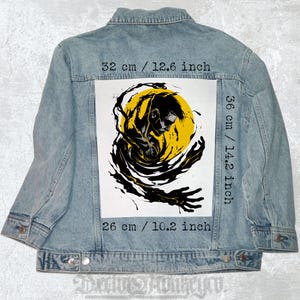 Pode incluir: Um patch de design gráfico preto e branco com uma pessoa com um círculo amarelo atrás. O patch tem 36 cm de altura e 26 cm de largura. O patch está em uma jaqueta jeans azul.