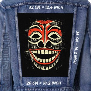 Può includere: Una giacca di jeans nera con una toppa di maschera tribale rossa, bianca e nera cucita sul retro. La toppa è larga 32 cm e alta 36 cm. Il testo "32 CM = 12.6 INCH", "36 CM = 14.2 INCH" e "26 CM = 10.2 INCH" è stampato sulla giacca.