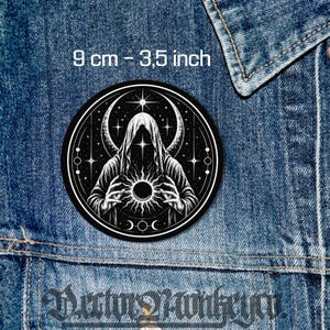 Pode incluir: Um emblema circular em preto e branco com uma figura encapuzada segurando um sol radiante, contra um céu estrelado com uma lua crescente. O emblema mede 9 cm de diâmetro e está sobre um fundo de tecido jeans azul.