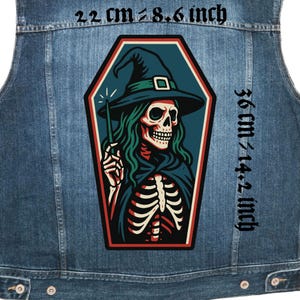 Puede incluir: Un chaleco vaquero azul con un parche grande en forma de ataúd que presenta una bruja esqueleto con cabello verde y un sombrero negro. El parche mide 22 cm de ancho y 36 cm de alto. La bruja sostiene una varita. El chaleco tiene botones metálicos.