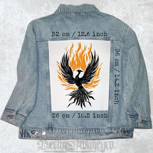 Könnte beinhalten: Hellblaue Jeansjacke mit einem schwarzen Phönix- und orangefarbenen Flammen-Design auf dem Rücken. Die Grafik ist 32 cm breit, 36 cm hoch und 26 cm von oben nach unten. Die Jacke hat einen klassischen Kragen und einen Knopfverschluss.